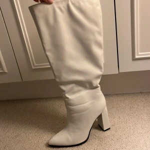 Vita höga stövlar med klack - Snygga vita stövlar med hög, bred klack och spetsig tå. Skorna är tillverkade i syntetmaterial med en slouchy passform som ger en trendig look. Perfekta för dig som vill sticka ut med ett par statement boots.