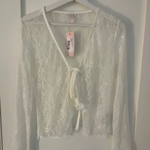 Vit spetstopp med knyt från Nelly - Superfin vit blus i spets från Nelly med långa ärmar och v-ringning. Blusen har knyt framtill och är genombruten med blommigt spetsmönster. Perfekt att styla med jeans eller kjol för en trendig look. Helt ny med prislapp kvar💕