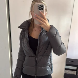 Grå pufferjacka från Polo Ralph Lauren - Snygg grå pufferjacka från Polo Ralph Lauren med hög krage och dragkedja framtill. Jackan har quiltad design, två sidofickor med dragkedja och Polo-logga på ärmen. Perfekt för kyliga dagar och riktigt skön att bära. Små små skador efter att den används