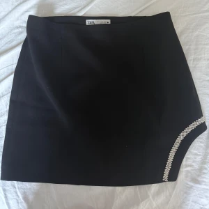 Svart kortkjol med strass från Zara - Svart kortkjol från Zara i stilren modell med en snygg strassdetalj längs ena nederkanten. Kjolen har en diskret dragkedja i sidan och är perfekt för dig som gillar en clean look med lite extra bling.
