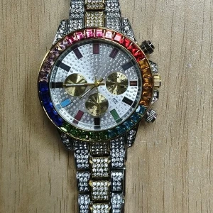 Färgglad blingad klocka med stenar - Säljer en riktigt maxad klocka med glittrande stenar i regnbågens färger runt urtavlan. Armbandet är i silver och guld med massor av små stenar. Urtavlan har tre mindre visare och datumvisning. Perfekt för dig som vill sticka ut och älskar bling!