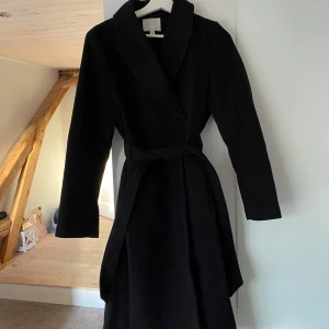 Svart kappa med bälte och krage - Stilren svart kappa med klassisk krage och knytskärp i midjan. Perfekt för dig som gillar minimalistisk stil och vill ha en jacka som funkar till många outfits. Perfekt inför höst/vintern. Den är använd en säsong så den är i väldigt bra skick!