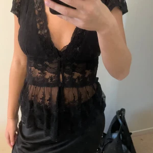 Svart spetsblus med knappar - Supersnygg svart blus med transparent spets och broderade detaljer. Djup v-ringning, korta ärmar och knappar framtill. Volang nertill. Har en spetts bh under!