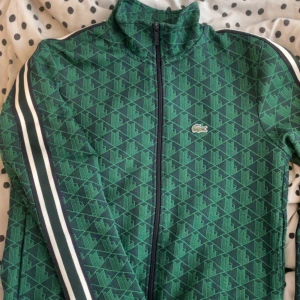 Grön mönstrad ziptröja från Lacoste - Säljer en grön ziptröja från Lacoste med geometriskt mönster och vita ränder längs ärmarna. Tröjan har hög krage, hel dragkedja och den klassiska krokodilloggan på bröstet. Regular fit och sportig vibe, perfekt för dig som gillar streetstyle.