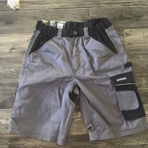 Grå arbets-shorts från Parkside - Säljer ett par grå arbets-shorts från Parkside med svarta detaljer och flera praktiska fickor. Shortsen har elastisk midja, bälteshällor och är tillverkade i bomull för extra komfort. Perfekta för dig som vill ha funktionella och slitstarka shorts till jobbet eller projekt.