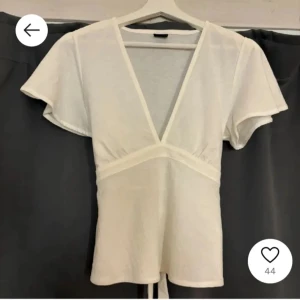 Vit omlottopp med v-ringning - Säljer en fräsch vit omlottopp med djup v-ringning och korta, luftiga ärmar. Toppen har en markerad midja och knytband baktill för en snygg siluett. Perfekt att styla med jeans eller kjol för en clean och trendig look.