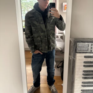 Camouflage overshirt med patches - Säljer en grön camouflage overshirt med coola US Airman-patchar och militärinspirerade detaljer på ärmarna. Jackan har flera fickor framtill och är tillverkad i bomull. Perfekt för dig som vill ha en streetig och avslappnad stil.