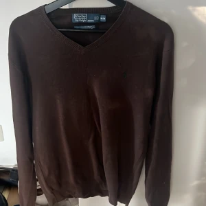 Brun v-ringad tröja Polo Ralph Lauren - Snygg brun v-ringad tröja från Polo Ralph Lauren i storlek M. Tröjan är tillverkad i 100% bomull och har ett broderat grönt Polo-logo på bröstet. Långärmad och med ribbade muddar vid hals, ärmslut och nederkant. Perfekt för dig som gillar klassisk stil.