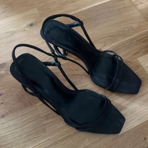 Svarta klackade sandaler från Zara - Svarta sandaler från Zara med smala remmar och fyrkantig tå. Skorna har en elegant hög klack och minimalistisk design som ger en stilren vibe. Perfekta för dig som gillar trendiga och enkla detaljer. Materialet är syntet och de har öppen tå.