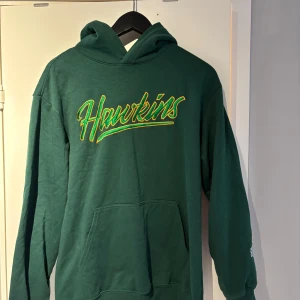 Grön Stranger Things hoodie Hawkins - Säljer en oversized grön hoodie med stor Hawkins-text broderad i gult och grönt på bröstet och Stranger Things-logga på ärmen.