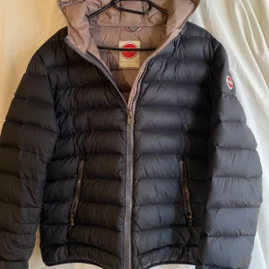 Svart dunjacka från Colmar - Snygg svart dunjacka från Colmar med huva och dragkedja framtill. Jackan har två fickor med dragkedja och quiltad design. Perfekt för kalla dagar och har en stilren look med diskret logga på ärmen. Vänster ficka är dragkedjan trasig så går inte o stänga annars jätte fin jacka inga hål
