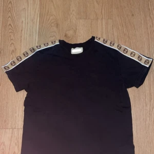Svart G.G t-shirt med logoband - Svart t-shirt från G.G med korta ärmar och klassisk passform. På axlarna finns beige band med bruna G.G-loggor som ger en lyxig och sportig vibe. Tillverkad i mjuk bomull som känns skön mot huden. Perfekt för dig som vill sticka ut med en ikonisk detalj.
