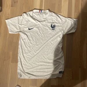 Frankrike Nike vit fotbollströja - Säljer en vit Frankrike fotbollströja från Nike med korta ärmar. Tröjan har små diskreta mönster i blått och rött över hela tyget, FFF-logga och Nike-swoosh på bröstet samt en trikolor-detalj i nacken. Materialet är lätt och andas, perfekt för match eller träning.