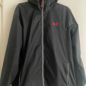 Svart vindjacka från Under Armour - Svart vindjacka från Under Armour med röd logga på bröstet. Jackan har huva, hel dragkedja framtill och två fickor med dragkedja. Tillverkad i lätt syntetmaterial, perfekt för blåsiga dagar. Enkel och stilren design med sportig känsla. Pris kan diskuteras 