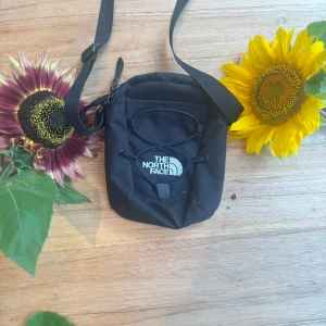 Svart axelväska från The North Face - Snygg svart axelväska från The North Face med justerbar rem och dragkedja baktill. Väskan har en praktisk frontficka med snörningsdetalj och tydlig logga framtill. Perfekt storlek för mobil, plånbok och andra småsaker. Tillverkad i slitstark polyester.