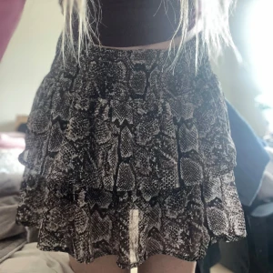 Kort kjol med ormmönster och volanger - Supersnygg kort kjol med volanger och elastisk midja. Kjolen har ett coolt ormmönster i svart, grått och beige. Lätt och flowigt material som ger en skön vibe. Perfekt för dig som vill sticka ut lite extra.