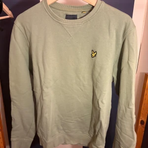 Grön sweatshirt från Lyle & Scott - Snygg ljusgrön sweatshirt från Lyle & Scott med klassisk rund hals och broderad gul logga på bröstet. Tröjan har långa ärmar och ribbade muddar vid ärmslut och nederkant. Perfekt för en chill och stilren look.