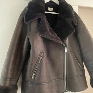 Brun teddyjacka shearling från Kappahl - Säljer en brun teddyjacka från Kappahl med stora slag, mjuk fuskpäls på insidan och detaljerade dragkedjor på fickor och framtill. Jackan har en loose passform och är perfekt för kyliga dagar. Snygg och trendig look med bred krage och markerade sömmar.