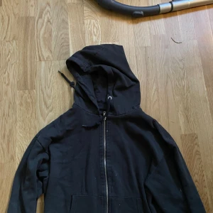 Svart hoodie med dragkedja - Svart hoodie med huva och snörning, hel dragkedja framtill och två fickor. Klassisk modell i mjuk bomull, perfekt för en avslappnad stil. Långärmad och enkel att matcha med det mesta.