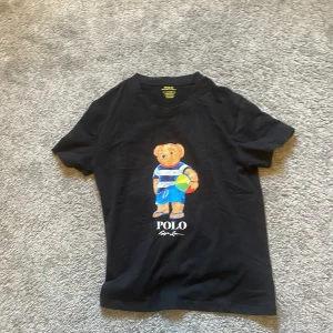 Svart Polo Ralph Lauren t-shirt med björn - Svart t-shirt från Polo Ralph Lauren med en färgglad björn på framsidan som håller en boll och har blå shorts. Modellen är custom slim fit med rund hals och korta ärmar. Materialet är mjuk bomull och trycket ger en chill vibe.