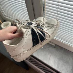 Valentino Rockstud vita sneakers - Snygga vita Valentino Rockstud sneakers med svart band och nitar på sidorna. Skorna har snörning, platt sula och är tillverkade i skinn. Väl använda därav priset. Torkade snabbt av innan bilderna togs men går säkert att fräscha till ännu mer med en rejäl rengörning och byte av skosnören!💗