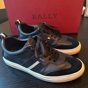 Svarta Bally sneakers med vit rand - Snygga svarta sneakers från Bally med vit rand på sidan och matchande svart snörning. Skorna har en rund tå, platt vit sula och detaljer i både skinn och mocka. Diskret Bally-logga på sidan och bakre hälflik med randigt band. Perfekt för dig som gillar stilrena och exklusiva sneakers.