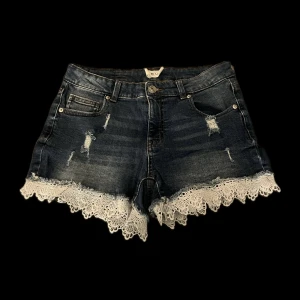 Mörkblå jeansshorts med spetskant - Snygga mörkblå jeansshorts från newyorker, med slitna detaljer och vit spetskant längst ner. Klassisk femficksmodell med knapp och dragkedja framtill. Perfekt för dig som vill ha en cool och feminin stil på sommaren. 
