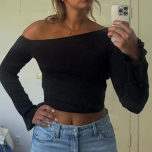 Svart offshoulder topp  - Säljer en svart ribbad offshoulder topp från NA-KD x Hanna Schönberg. Toppen är croppad med långa vida ärmar och har en snygg, figurnära passform. 
