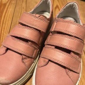 Rosa sneakers från Ecco i skinn - Snygga rosa sneakers från Ecco i mjukt skinn med tre breda kardborreband för enkel påtagning. Skorna har vit sula och rund tå, perfekt för en avslappnad och trendig look. Passar dig som gillar stilrena och bekväma skor med lite färg.