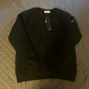 Svart sweatshirt från Stone Island - Svart sweatshirt från Stone Island med klassisk rund hals och ribbade muddar. Tröjan har den ikoniska logotypen på vänster ärm och är gjord i mjuk bomull. Perfekt för dig som gillar stilrena och enkla plagg med streetkänsla.