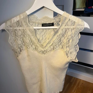 Gräddvit spetstopp från Fashion, Storlek S - Supersöt gräddvit topp med breda spetsdetaljer upptill och v-ringning från Fashion i storlek One Size, passar bäst på  en S🫶 Toppen är ribbad nedtill och har ärmlös design med romantisk känsla. Använd ett fåtal gånger och i mycket bra skick🤍 Perfekt till både vardag och fest! 