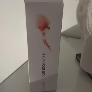 Kenzo Amour parfym 100ml - Kenzo Amour parfym i en elegant, mörkrosa flaska med smal, böjd form. Flaskan är tillverkad i plast eller glas och har en modern, stilren design. Doften är känd för sina mjuka, blommiga och varma toner. Kommer i originalförpackning med konstnärlig design.