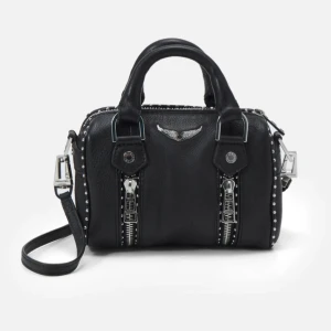 Svart handväska från Zadig & Voltaire - Cool svart handväska i skinn från Zadig & Voltaire med silverfärgade detaljer, dubbla dragkedjor framtill och nitar längs kanterna. Väskan har både handtag och axelrem, samt en ikonisk vinglogga i metall framtill. Perfekt för att lyfta vilken outfit som helst.