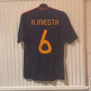 Spaniens VM-tröja Iniesta 2010 Adidas - Säljer en officiell Adidas Spanien fotbollströja från VM-finalen 2010 med Iniesta 6 på ryggen. Tröjan är mörkblå med gula och röda detaljer, broderat landslagsemblem, matchinfo på bröstet och VM-märke på ärmen. Tillverkad i lätt funktionsmaterial, perfekt för samlare eller fans.