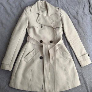 Beige trenchcoat från Forever 21 - Snygg beige trenchcoat från Forever 21 med klassisk dubbelknäppning, bred krage och bälte i midjan. Jackan har långa ärmar, två fickor framtill och stilrena knappar. Kappan är som ny och säljs på grund av att den är för liten. 