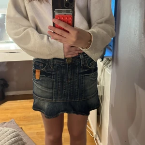 Mörk jeanskjol med volangkant - Säljer en snygg mörkblå jeanskjol med slitningar, volangkant och coola bakfickor. Kjolen har en dekorativ patch med stenar bak och en orange detalj vid fickan fram. Klassisk knapp och dragkedja framtill. Perfekt för dig som gillar streetstyle.