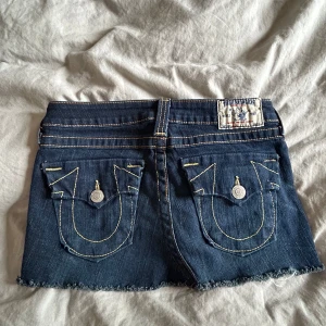 True Religion jeanskjol mörkblå - Mörkblå jeansshorts från True Religion med ikoniska bakfickor och kontrastsömmar. Rå kant nertill för en edgy vibe och klassiska detaljer som knappar och fickor fram och bak. Perfekta för sommaren och ger en cool streetstyle-look.
