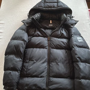 Svart pufferjacka från BOSS med huva - Säljer en svart pufferjacka från BOSS med huva och dragkedja. Jackan har en clean look med breda, quiltade sektioner och diskret BOSS-logga på ärmen. Perfekt för kalla dagar och riktigt snygg till streetwear-stilen. Med avtagbar luva. Ordinarie pris 3600kr.