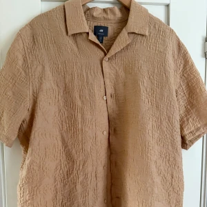 Beige kortärmad skjorta från H&M - Säljer en beige kortärmad skjorta från H&M i regular fit. Skjortan har en krinklad struktur, klassisk krage och knäppning framtill. Perfekt för dig som gillar en avslappnad men ändå stilren look.