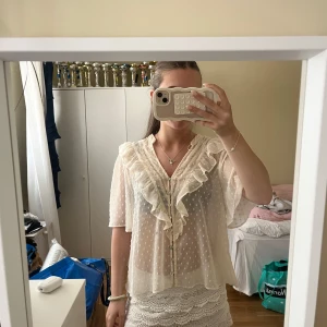 Vit blus med volang - Supersöt vit blus i transparent tyg med små prickar, volang längs v-ringningen och knappar framtill. Blusen har korta, vida ärmar och romantisk känsla. Perfekt för dig som gillar en feminin och luftig stil.