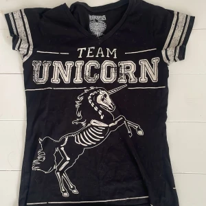Svart Team Unicorn t-shirt från Heartless - Svart t-shirt från Heartless Clothing med coolt skelett-unicorn-tryck och texten 'Team Unicorn' både fram och bak. V-ringad modell med korta ärmar och sportiga ränder på ärmarna. Perfekt för dig som gillar alternativ stil och unika prints.