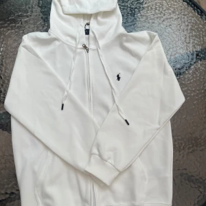 Vit hoodie från Polo Ralph Lauren - Snygg vit hoodie från Polo Ralph Lauren med dragkedja och huva. Klassisk broderad logga på bröstet i svart. Tröjan har långa ärmar, snörning i huvan och ribbade muddar. Perfekt för en clean och stilren look.