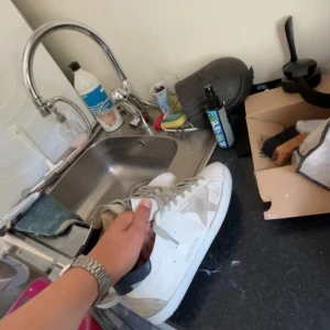 Vita sneakers från Golden Goose - Säljer ett par klassiska vita Golden Goose sneakers med grå stjärndetalj på sidan och grå snörning. Skorna har en rund tå och är tillverkade i skinn med slitna detaljer för en cool, vintage look. Perfekta för dig som gillar streetstyle och vill ha något unikt. Dom är lite sönder för ramla med scooter som man ser men har täckt med whiting cream allt og får man med