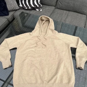 Sand merinoull hoodie  - Beige stickad hoodie med huva och snörning. Den är i färgen sand och har en tajt passform. Perfekt för dig som en enkel och stilren look. Passar till det mesta och är riktigt skön att ha på sig 