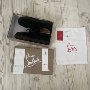 Christian Louboutin svarta sneakers med nitar - Säljer ett par svarta sneakers från Christian Louboutin med ikoniska röda sulor och coola svarta nitar på tån. Skorna har snörning och är tillverkade i mocka med läderdetaljer. Perfekta för dig som vill sticka ut med en lyxig och edgy stil.