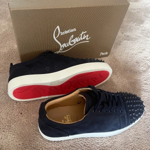 Christian Louboutin svarta mockasneakers - Snygga svarta sneakers från Christian Louboutin i mocka med ikoniska röda sulor och coola nitar på tån. Skorna har snörning och vit platt sula, perfekt för dig som vill sticka ut med en lyxig och edgy stil.