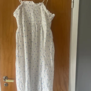 Vit blommig långklänning med smock - Supersöt långklänning i vitt med små blå blommor, smock upptill och tunna axelband. Volangdetalj vid bysten och luftig passform, perfekt för varma dagar. Lätt och skönt material som känns som bomull.