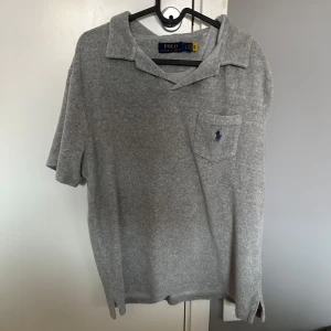 Grå piké från Polo Ralph Lauren - Klassisk grå pikétröja från Polo Ralph Lauren med korta ärmar och bröstficka med broderad blå logga. Mjuk bomullskänsla och avslappnad passform, perfekt till jeans eller shorts. Snygg krage och diskret slits i sidorna. används fåtal gånger 