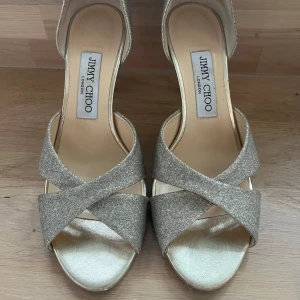 Jimmy Choo klackar - Snygga klackar från Jimmy Choo i guldigt glittermaterial med korsade band framtill och öppen tå. 