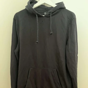 Svart hoodie med huva och ficka - En klassisk svart hoodie med huva och dragsko, samt en stor magficka framtill. Perfekt för chill dagar eller när du vill ha en enkel och stilren look. Långärmad och i mjukt bomullsmaterial.